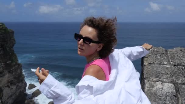 海に巻き茶色の髪と眼鏡を持つ若い白人女性のスローモーション 女性は高さから海の自然を楽しむ 暖かい国で休暇中の魅力的な無料の女性 自由幸福概念 — ストック動画