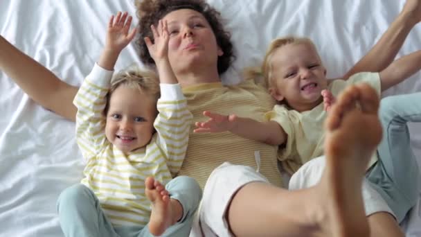 トップ表示スローモーションベッドの上の子供たちと幸せな陽気な女性たちの足を笑って遊んで笑顔をぶら下げている 家族との幸せな週末の休日 ベッドの上の女性と子供は彼らの足を危険にさらす楽しい — ストック動画