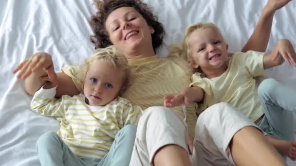 带着两个女儿躺在床上的快乐有趣的女人的头像 一个女人 带着女儿笑着摇动着腿 胳膊在床上跳舞 动作缓慢 幸福童年和母亲的概念 — 图库视频影像