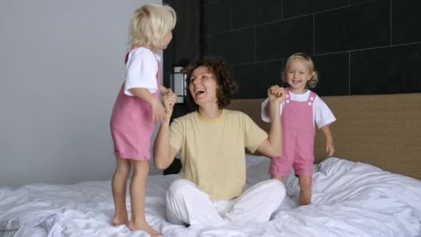 二人の双子の女の子は 楽しさと人生を楽しんで 若い魅力的な白人の母親とベッドの上でジャンプします ママと子供たちは休みの日にベッドで楽しんでいます 娘の女の子はベッドの上でジャンプ ママの手を保持 — ストック動画