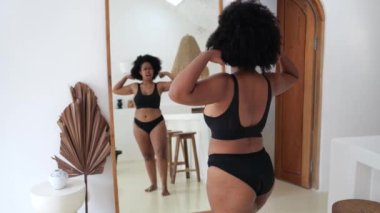 Çekici, siyah, kilolu, iç çamaşırlı bir kadın aynanın önünde gösteriş yapıyor ve güzel vücuduna hayran. Siyah vücudu ile uyumlu gururlu bir kadın. Görünüşün için pozitif ve sevgi 