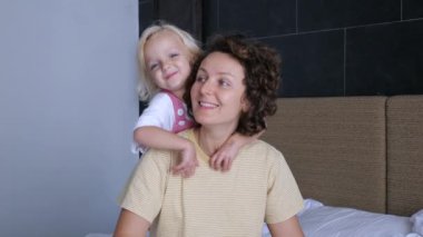 Sevimli beyaz anne ve çocuğu sevgiyle kucaklıyor, nazikçe gülümsüyor ve gülüyor. Sevgi, şefkat, anne ve kız bebek arasındaki duygusallık. Kız çocuğu annesinin boynuna arkadan sarılıyor.