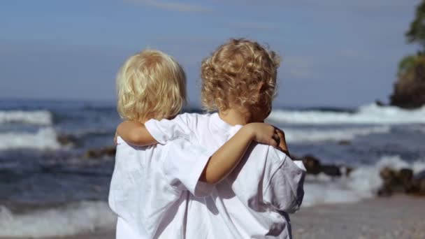 两个孩子在海滨或海里肩并肩抱在一起的后视镜 两个孩子在岸上拥抱 享受着吃棒棒糖的美景 小高加索孩子从海上旅行中获得了新的印象 — 图库视频影像