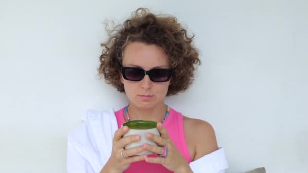 黒眼鏡の魅力的な白人の若い女性は外に座っている間に緑色のスムージーを楽しんでいます カップの中で栄養価の高いスムージーや抹茶を減量するための適切な栄養 おいしいお酒を楽しむ — ストック動画
