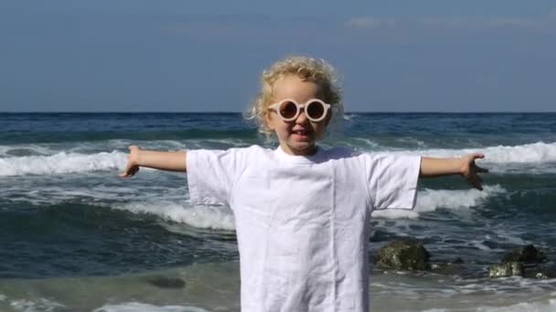 海の波の海岸洗浄の背景に小さな巻きブロンド白人の女の子 ファッショナブルな女の子は彼女の腕を伸ばして太陽の下で立ち 笑顔で残りの休日を楽しんでいます 暖かい海での週末 — ストック動画