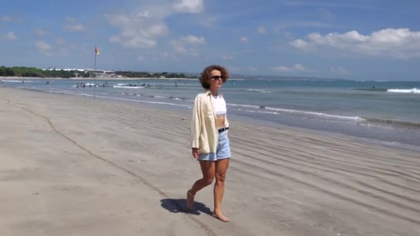 女性は晴れた日にビーチを歩き 彼女の手で彼女の巻き毛をまっすぐにします 海のビーチに沿って歩くことはリラックスして癒しです コンセプト心の健康を維持するためにビーチ沿いを歩く — ストック動画