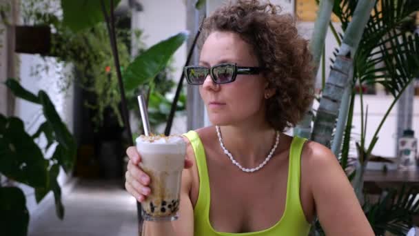靠近点一个戴太阳镜的金发女人拿着一杯泡茶 在餐馆里用奶油和泡沫重新装饰泡茶 在暑期餐厅享受生活 喝泡泡茶 — 图库视频影像