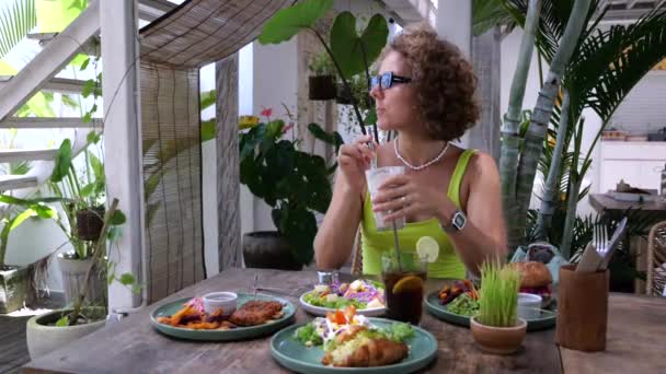 サングラスのブロンドの女性は 飲み物を攪拌し 夏のレストランで昼食を取っています 観光客のための夏のレストランメニュー バーガー クロワッサン フレンチフライ ビーフリブ ミルクシェイク 夏のレストランで休息 — ストック動画