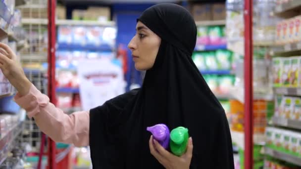 スーパーマーケットの店の棚に囲まれた女性は 個人的な衛生のために良い石鹸を探しています アルメニアのムスリム女性はラベルに構成を読み 液体石鹸を選択する セルフケアを維持する — ストック動画