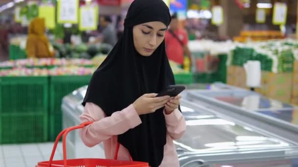 히잡의 무슬림 여성은 빨간색 바구니와 슈퍼마켓에 신속하게 그녀의 전화에 외국인 — 비디오
