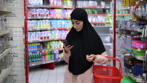 女性は店の棚の真ん中に赤いバスケットが付いているスーパーマーケットに立っている間スマートフォンを使ってメッセージを送ります ヒジャブのムスリム女性は 店で買い物をしながら 彼女の携帯電話で積極的に対応しています — ストック動画
