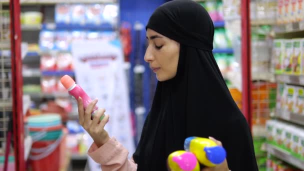 一名头戴头巾的穆斯林妇女站在那里 手里拿着三种化妆品 比较化妆品的成分和价格 一位健康的伊斯兰妇女为头发和身体选择合适的护肤产品 — 图库视频影像