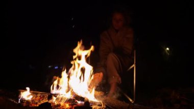 Geceleri kamp ateşinin yakınında, genç bir bayanla ateşin yanında. Ağır çekim video: Kamp yapan kadın sandalyeye oturur, battaniyeye sarılır ve ateşle ısınır. Kamp ateşinde çakmak kullan.
