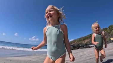 İki mutlu küçük sarışın kız yaz plajında kameraya doğru koşuyor. Mayo giymiş, gülen, güzel plajlarda koşan, eğlenmek için koşan anaokulu öğrencilerinin dinamik videosu.