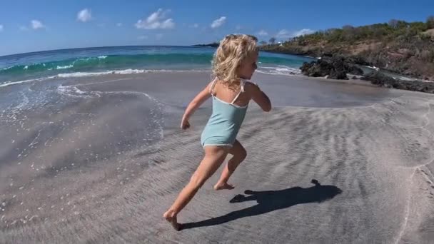 海岸沿いの湿った砂の上を走るエネルギッシュな少女の側面 ブロンドカーリーヘアガールのワイドショットは バックグラウンドランニングで荒れ狂う海に走る大きな石に実行され 海による休暇で素晴らしいスポーツです — ストック動画