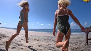 Mayo giymiş koşan iki mutlu kızın okyanusta yüzmeye gidişinin alt görüntüsü. Güneş pansiyonlarının ve güneş şemsiyelerinin önünden yüzmek için sıcak kumda koşan sevimli çocukların dinamik videosu. Yazın okyanusta yüzme kavramı