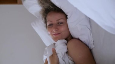 Mutlu genç bir kadının güzel, tembel bir hafta sonu uyanıp yumuşak yastığın üzerinde rahat bir yatakta yattığı dikey bir video. Dinamik çekim: yatakta yumuşak battaniyenin altında yatan sağlıklı bir kadın, gülümsüyor. Yatak konseptinde uzanmak