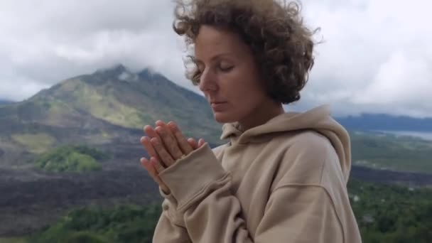 フーディーの女性が山で祈り 自然とつながり 風のヘアボウリングをしています ダイナミックな人形は 山で祈る精神的な瞬間のビデオをズーム 穏やかさ 山で祈る平和 — ストック動画