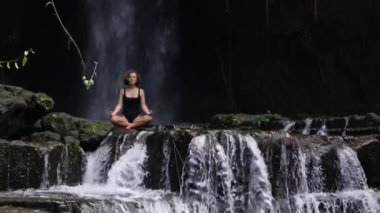 Şelalenin tepesinde mayo giymiş genç bir kadın büyük taşların üzerinde otururken meditasyon yapıyor. Şelalenin tepesinde büyük taşların üzerinde sakin bir şekilde oturan kadının sinematik görüntüsü. Şelalenin tepesindeki meditasyonu kabul et