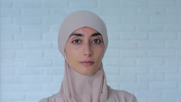 自给的穆斯林妇女的画像 头戴浅色头巾 看着相机 年轻的穆斯林妇女站在白砖墙的背景下进行特写 穆斯林妇女可以自给自足的概念 — 图库视频影像