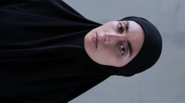 Müslüman kadının büyüleyici bakışlı dikey videosu kameraya bakıyor. Siyah tesettürlü genç müslümanların yakın çekimi gözlerini açtı ve kameraya baktı. Çekici Müslüman güzelliği kavramı. Koyu renk başörtüsü