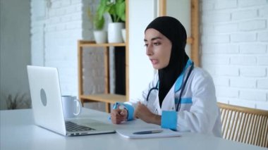 Arap kadın doktor dizüstü bilgisayar üzerinde çalışırken online danışmanlık yapıyor. Modern teknolojiler dünyanın herhangi bir yerinden uzaktan çalışmanızı sağlar. Kadın doktor kendini evinden uzakta çalışmaya adadı..