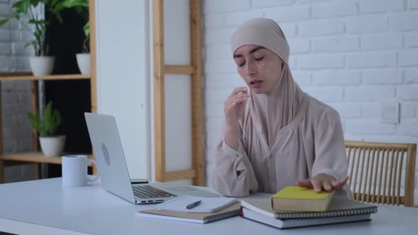 東洋の若い女性学生は学習プロセスに忙しい ヒジャーブのムスリム女性は本 ノートパソコンの後ろに座り 深刻な試験に備えています 新しい知識は女性にとって困難であり 緊張を引き起こす — ストック動画