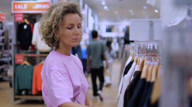 Giyim mağazasındaki kadın bluzlara bakıyor, alışveriş sürecinden zevk alıyor. Alışveriş vaatleri özellikle başarılı ve keyifli alışverişler için cömerttir. Alışveriş bağımlılığı ve alışveriş bağımlılığı kavramı