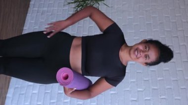 Eşofmanlı güçlü bir kadının yoga minderini tutarken çekilmiş dikey videosu. Gülümseyen kadın ev çalışmasından sonra enerji ve pozitiflikle doludur. Kadınlar için spor eğitimi kavramı