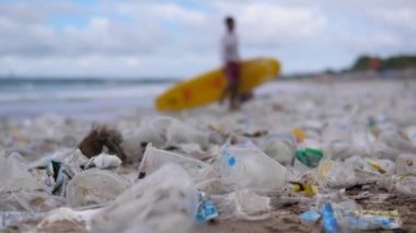 Plajda plastik kirliliği ve arka planda sörf tahtası olan bir adam. Plajdaki plastik çöplere yakın çekim çevre kirliliğine yol açacak. Plastik kirliliği ve çevre sorunları kavramı