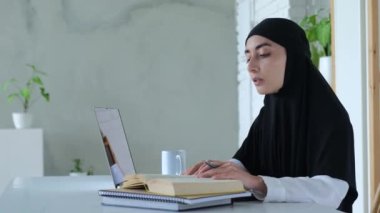 Siyah tesettürlü Müslüman kadın internette okuyor. Yan bakış: Müslüman kadın kitaptan bilgi yazdı, sonra da bilgisayarını internetten kullandı. Kavram: dizüstü bilgisayar, defterler ve kitaplar çevrimiçi bilgi edinmeye yardımcı olur
