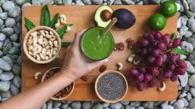 Sağlıklı beslenmek için tahta tahtaya yassı yeşil meyveler, sebzeler, fındıklar ve tohumlar serilmiş. Kadın, bardağı samanla doldurur. Yeşil smoothie ile sağlıklı sabah kahvaltısı
