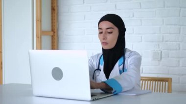 Müslüman kadın doktor dizüstü bilgisayarda çalışıyor. Yakın çekim: tesettürlü Müslüman kadın doktor dizüstü bilgisayar ekranına bakar ve deftere bir şeyler yazar. Konsept: Laptop başında oturan Müslüman kadın doktor