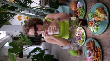 Dikey video kıvırcık sarışın bir kafede milkshake içiyor. Yazın turistler için en iyi tatlı milkshake 'tir. Yaz restoranı menüsü: hamburger, sığır pirzolası, avokado, kruvasan, patates kızartması, milkshake.