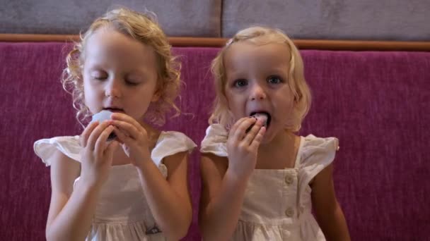 2人の双子の女の子がアーモンドマカロニクッキーを食べている ドレスに身を包んだ金髪の女の子たちは アーモンドマカロニクッキーを熱烈に食べている カフェでのダイナミックなショット かわいい子供たちはアーモンドマカロニクッキーを食べる — ストック動画