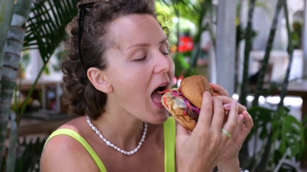 フレームを閉めろ 空腹のブロンドはハンバーガーの大きな部分を噛み 喜びで噛む 夏のトロピカルレストランで大きなジューシーなバーガー コンセプト おいしいバーガーを噛み あなたの空腹を満たす — ストック動画