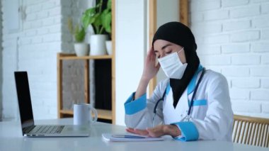 Müslüman doktor ağır ağır iç çekiyor, tıbbi maskeyi çıkarıyor ve tapınağı çok yoruyor. Masadaki Müslüman doktor kalemini bırakır, maskesini çıkarır ve alnını ve şakağını ovar. Kavram: yorgun Müslüman doktor