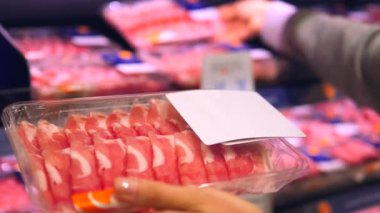 Süpermarkette kadın eti dikkatle seçer. Bir eliyle et paketini tutarken, diğeri raftaki çeşitli et ürünlerini inceliyor. Mağazadaki seçimler için geniş aralıklı etlere yakın çekim.