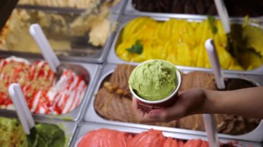 Kapalı çekimde, çeşitli tatlarla dolu tepsilerin karşısında renkli bir dondurma. Seçme tatları inanılmazdır, klasikten en olağanüstüye. Dondurmanın tadı unutulmaz bir zevktir..