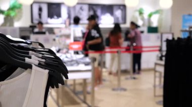 Kıyafet butiği. Seçici odak: Tişörtler önde, arka planda asılı, müşteriler seçtikleri kıyafetlerin parasını kasada ödüyorlar. Konsept: Butikte kıyafet almak uygun ve güvenlidir