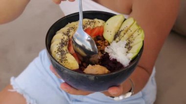 Meyveli meyve suyu kasesi. Yakın plan: muz, armut ve çilek parçaları üzerine susam tohumları serpiştirilmiş meyve suyu kasesi. Kadın eli kaşıkla granola alır. Meyve suyu kaseli sağlıklı beslenme konsepti