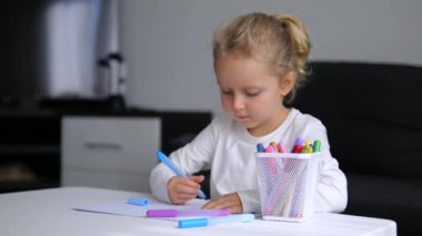 Sarı kıvırcık saçlı güzel kız masaya oturur ve keçeli kalemlerle renkli resimler çizer. Çocuk kendini yaratıcı süreçlere kaptırıyor. Sevimli bebek çiziyor, kağıt üzerinde büyülü bir dünya yaratıyor..