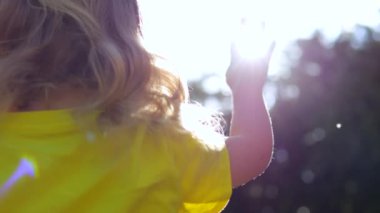 Sarı tişörtlü küçük kız, merak dolu, yumuşak yaz güneşine doğru ellerini uzatıyor. Parlak ışınlar yaprakları delip geçer ve narin parmakları delip geçer Kız dünyayı merakla dolaşır