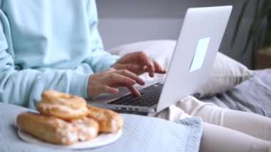 Kadın, yanında bir fincan kahve ve bir tabak fırınla dizüstü bilgisayarla yatakta sessiz vakit geçirmekten hoşlanıyor. Kadın rahat ev hayatının yanında dizüstü bilgisayarlı bir fincan kahveyle yatakta oturuyor.