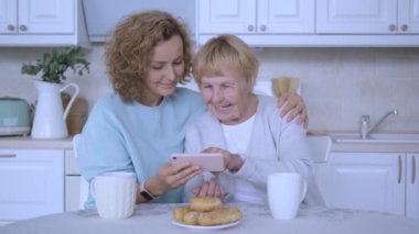 Büyükanne ve torun mutfak masasında kahve fincanları ve lezzetli pastalarla oturuyorlar. Torun, büyükannesine cep telefonunu nasıl kullanacağını öğretiyor. Nesiller arasında güçlü bir aile bağı.