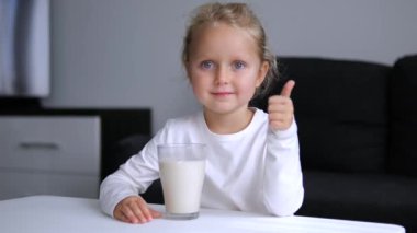 Küçük tatlı kız, yanında bir bardak sütle masada oturmuş kameranın beslenme gelişimine bakıyor. Beyaz tenli küçük bir kız, masa başında oturmuş kameraya besleyici yiyecek içeceklerine bakıyor.