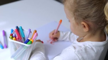 Kız evde kalemle hevesle çizim yapıyor. Kız kâğıt üzerinde parlak bir resim yaratıyor. Yaratıcı kız, çok renkli keçeli kalemleri kağıt üzerinde özenle hareket ettiriyor. Genç ressam hayal gücünü geliştiriyor.