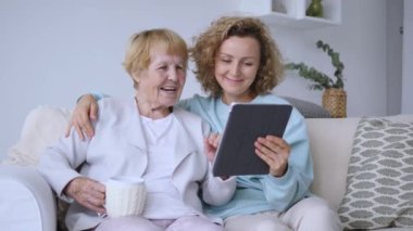 Büyük anne ve büyük kız kanepeye oturmuş tabletteki resimlere bakarken mutlu aile ilişkilerine bakıyorlardı. Emekli anne, yetişkin kız kanepeye oturmuş mutluluğu paylaşıyor.