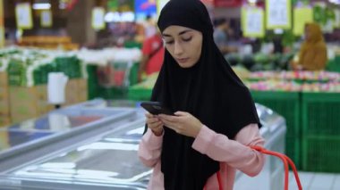 Süpermarketteki Müslüman kadın alışveriş arabası tutarken aktif olarak telefon kullanıyor. Modern teknoloji ve gelenek süpermarkette birleşerek, süpermarkette alışveriş ve çevrimiçi iletişimi kolayca birleştirir..