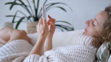 Sıcak elbiseli bir kadın kanepede dinlenmeyi, telefonda sosyal ağlarda gezinmeyi seviyor. Koltukta poz vermek tamamen rahatlamayı yansıtır. Anlık rahatlama, ev ve modern teknolojinin rahatlığını birleştirir..